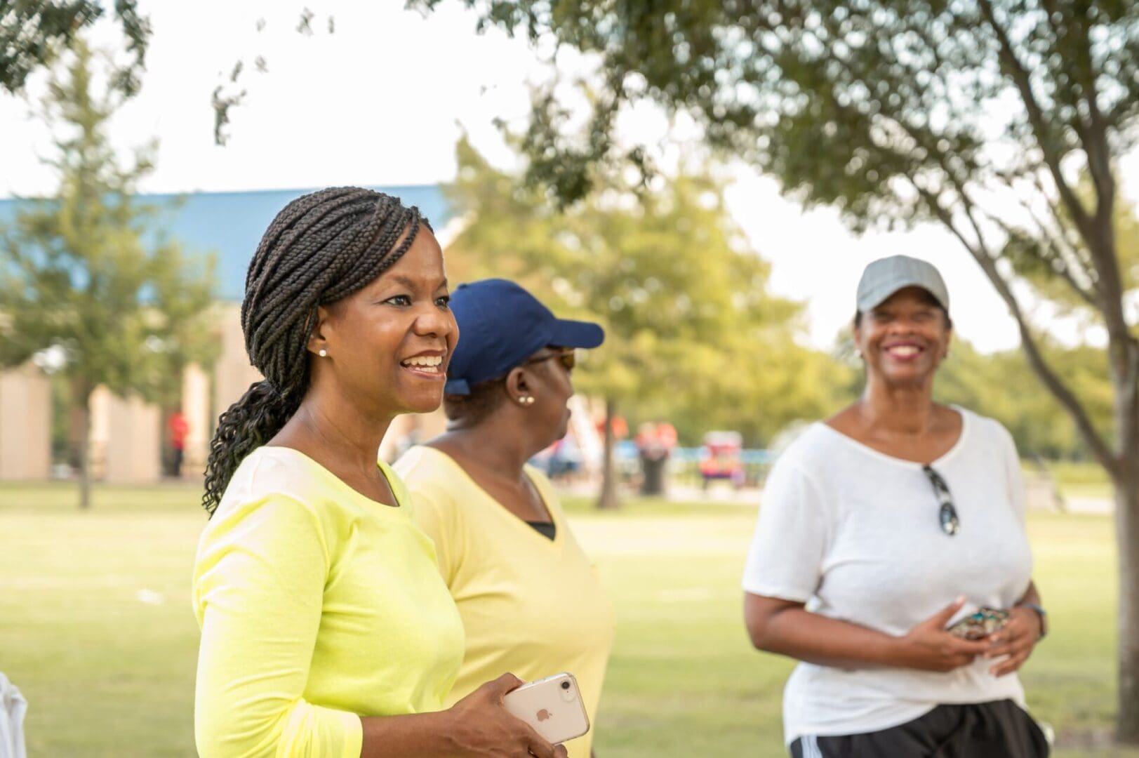 Three women are standing in a park and smiling.